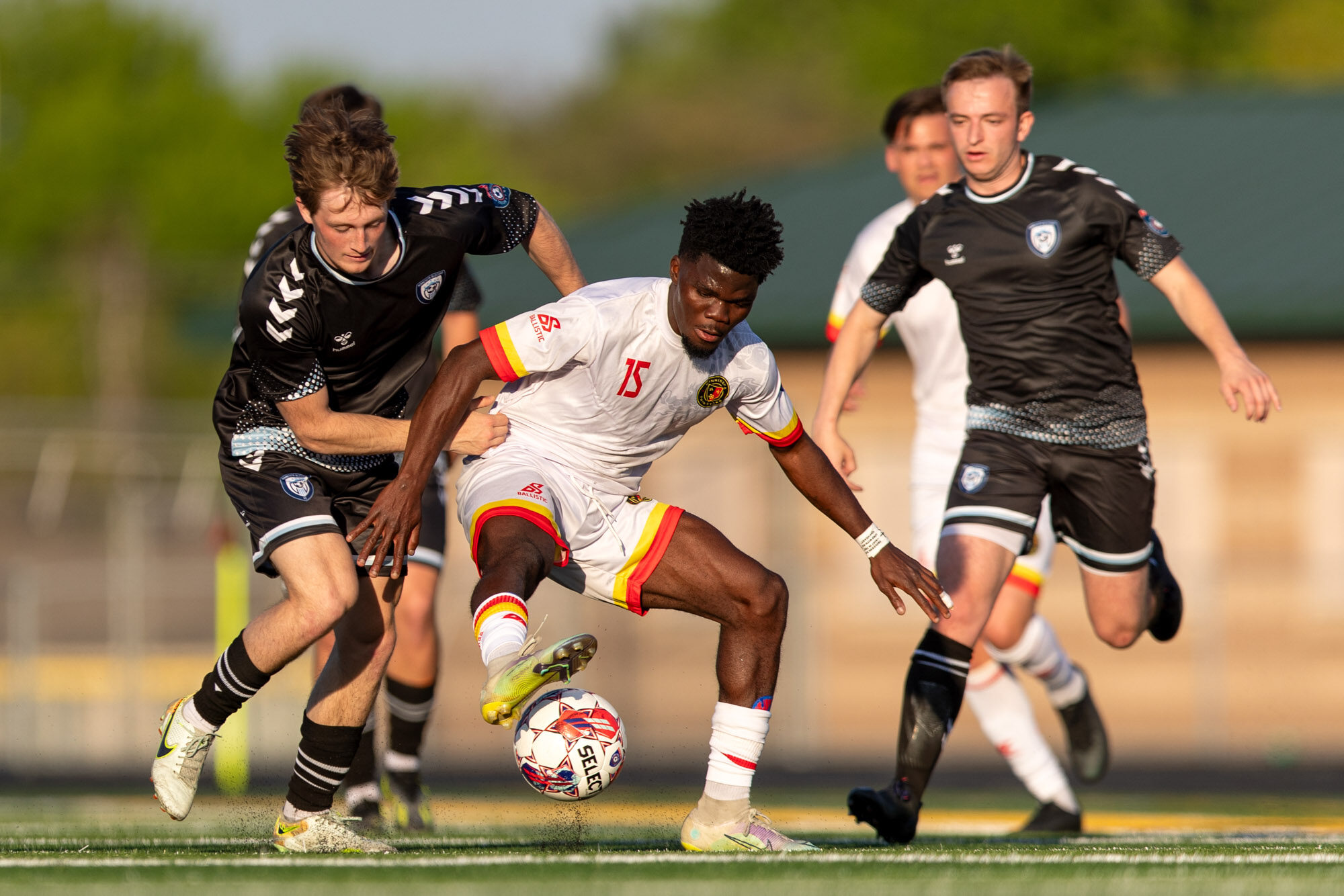 Two white soccer players in black kits run either side of a black man in a white kit, who is controlling the football