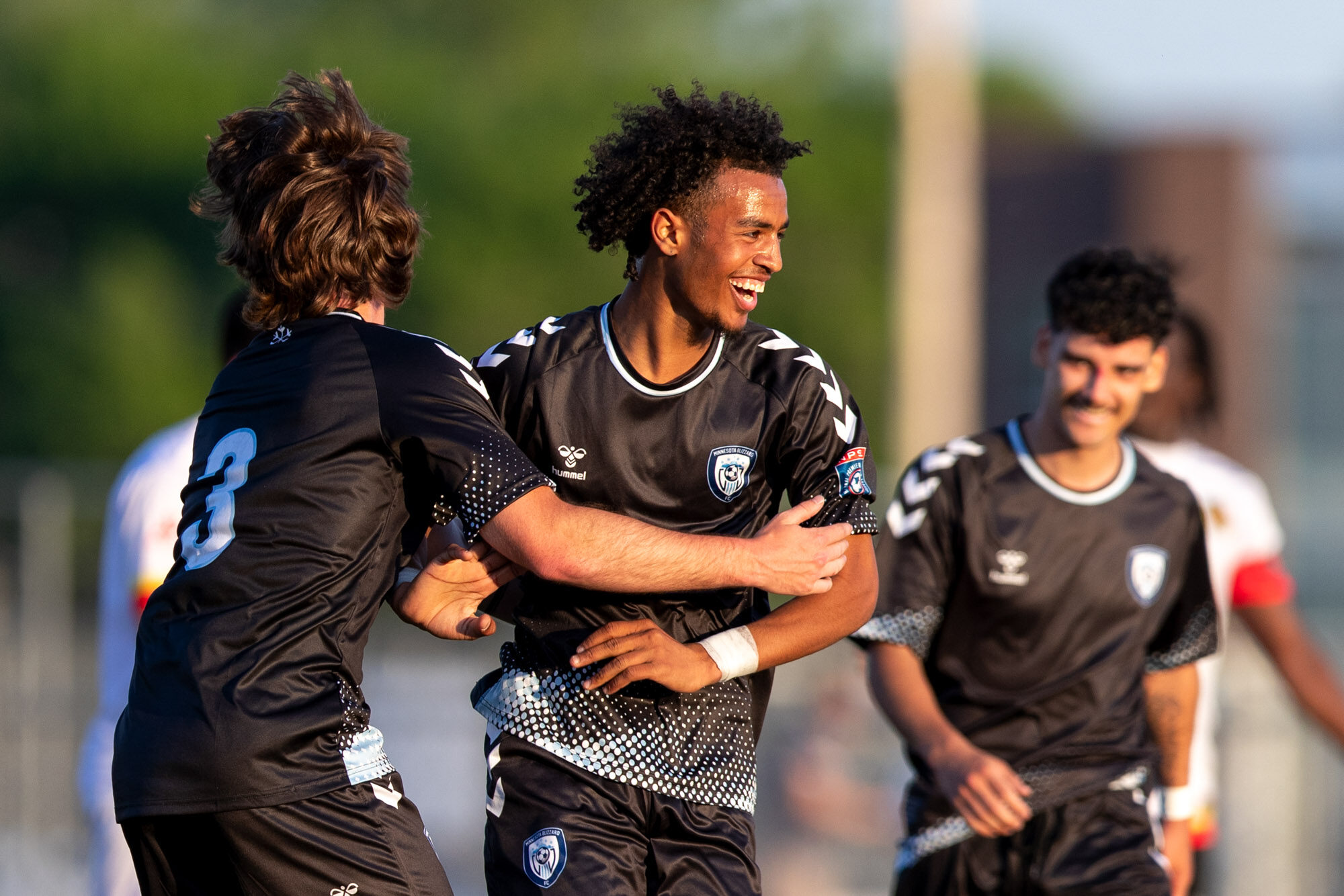 Three soccer players in all black kit celebrate. The central figure is a black man facing the camera with a broad smile. 