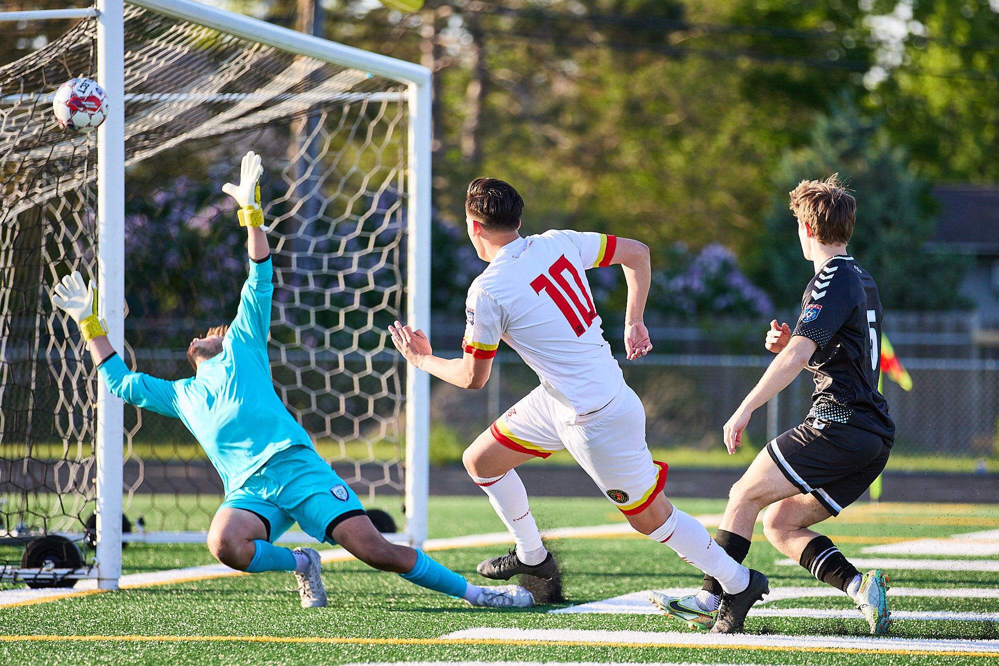 A turquoise-kitted goalkeeper dives upward, the ball just above his reach and next to the near post. A white kitted man wearing 10 watches on, having made the shot. A defender in a black kit looks on. 