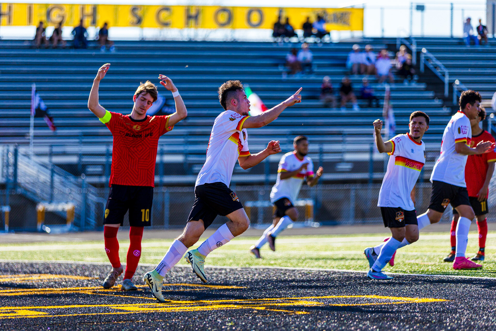 Red shirted soccer player with arms aloft in frustration. White shirted player center-frame pointing to the right