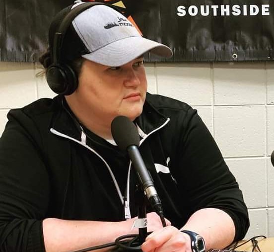Bridget sits behind a microphone wearing headphones, grey ball cap, and black Puma track jacket (on brand).