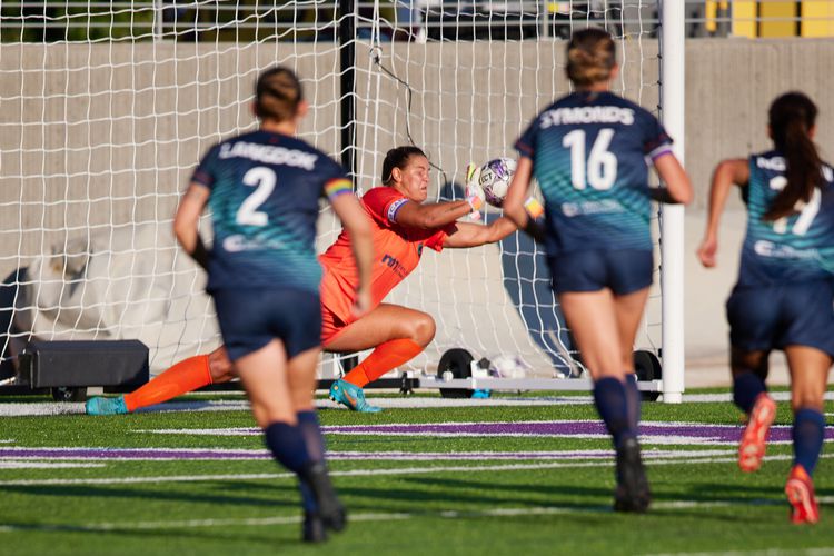 Goalkeeper Sarah Fuller makes a diving save in goal, framed between teammates Langdok and Symonds.
