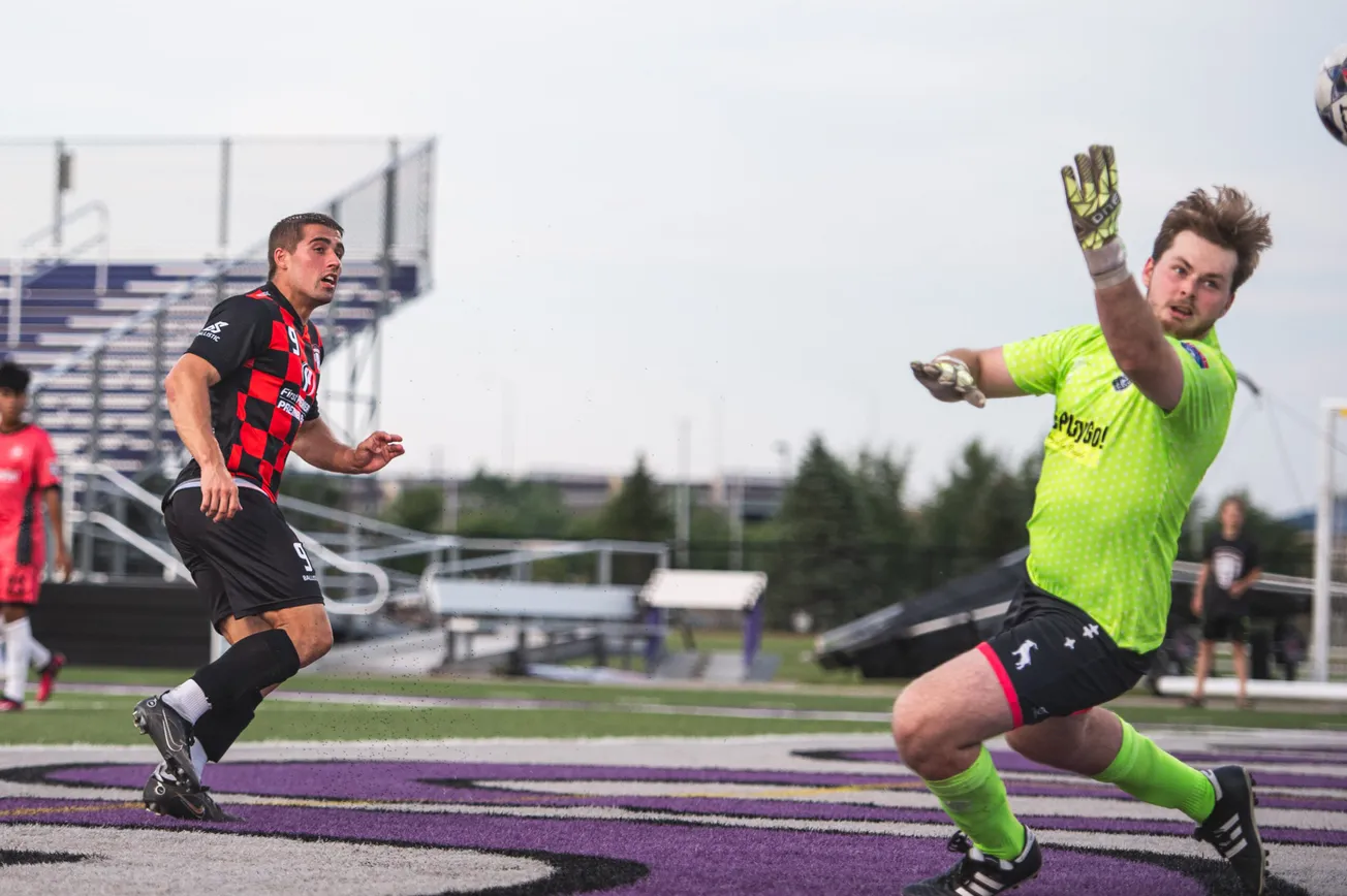 A man in a black and red check jersey and black shorts is in left of the frame, looking from left to right. A goalkeeper in a