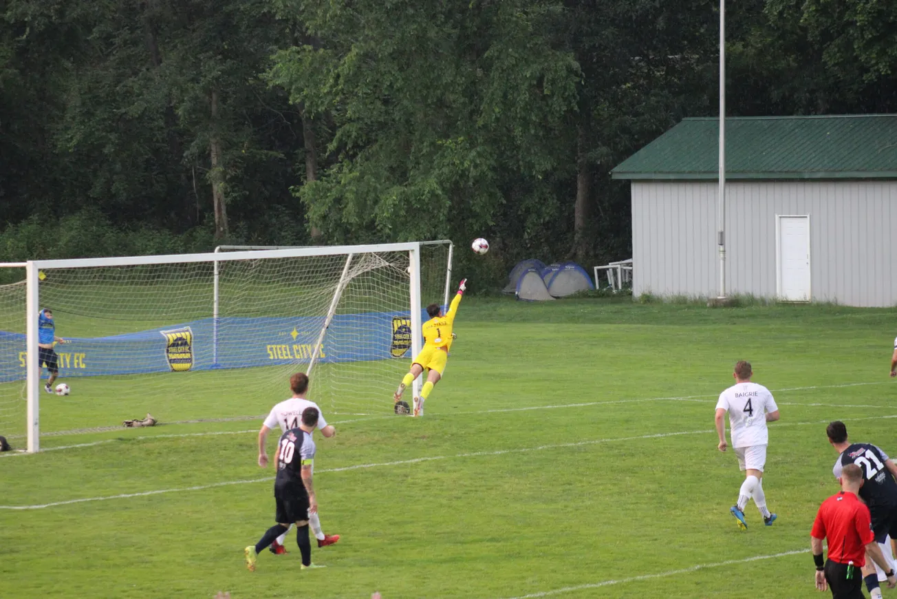 A yellow kitted goalkeeper is diving diagonally to the right, the ball is just above his left hand