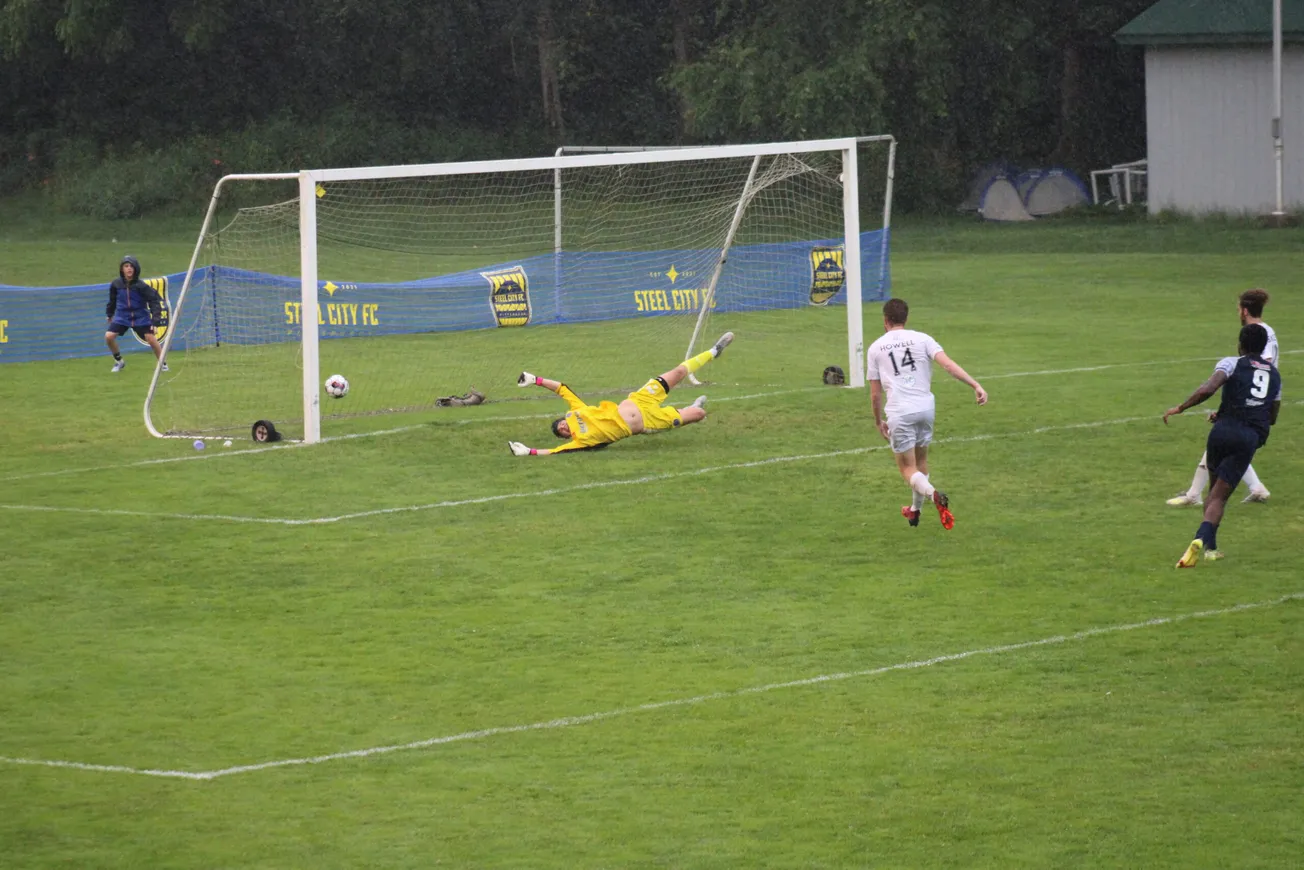 The ball is in the center-left of the frame, just inside the goal. Yellow kitted goalkeeper is horizontal across the goal. Wh