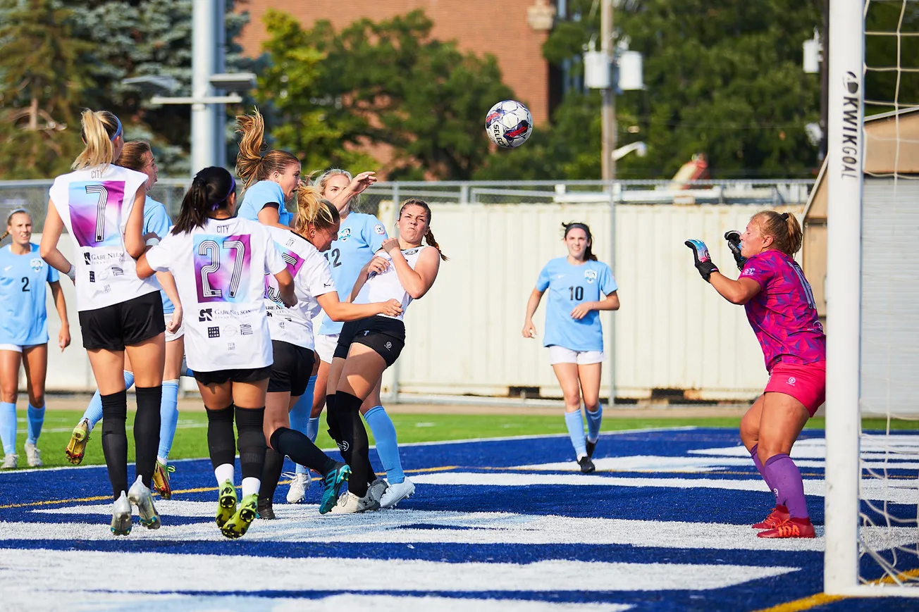 A group of women in soccer kits fight for a ball, which is in the middle of the frame. A goalkeeper is pictured in the right