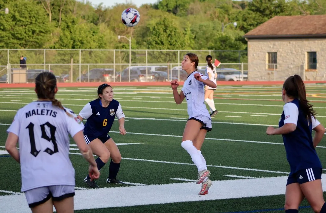 Left of frame, Alexa Maletis 19, with her back to the camera. Center frame: two players contest an aerial ball (one in navy k
