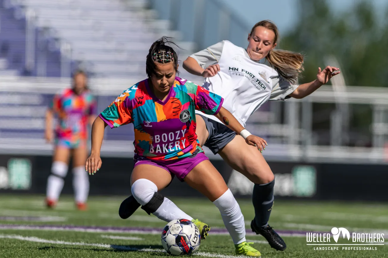 A woman with multicolored beads in her hair and a multicolored soccer jersey in foreground, soccer player in white jersey in