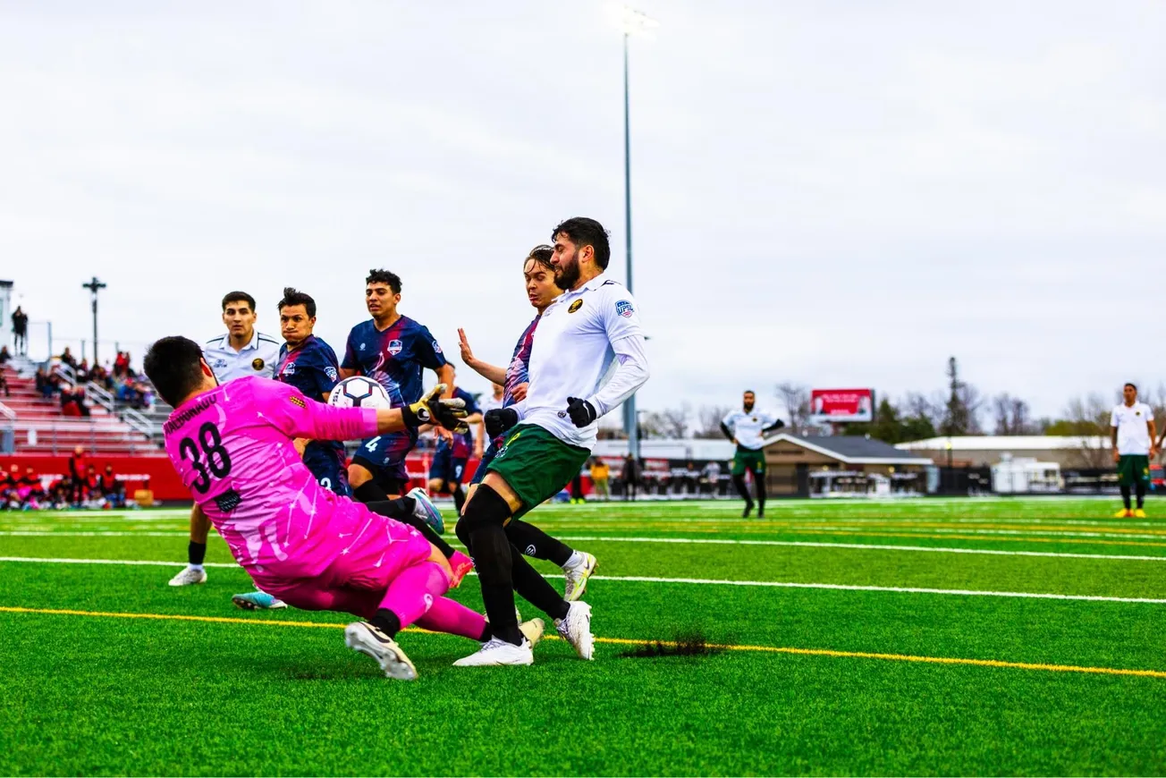 A pink shirted goalkeeper in the center of the frame stops a football with 5 other players looking on