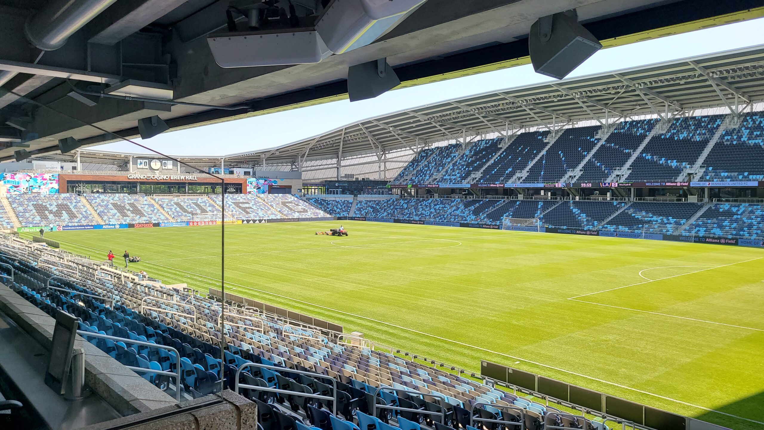 Allianz Field interior from the Southwest corner of the concourse: a lawn care tractor tends to the grass. The seats are empty. The Brew Hall and manual clock/scoreboard are in the distance.